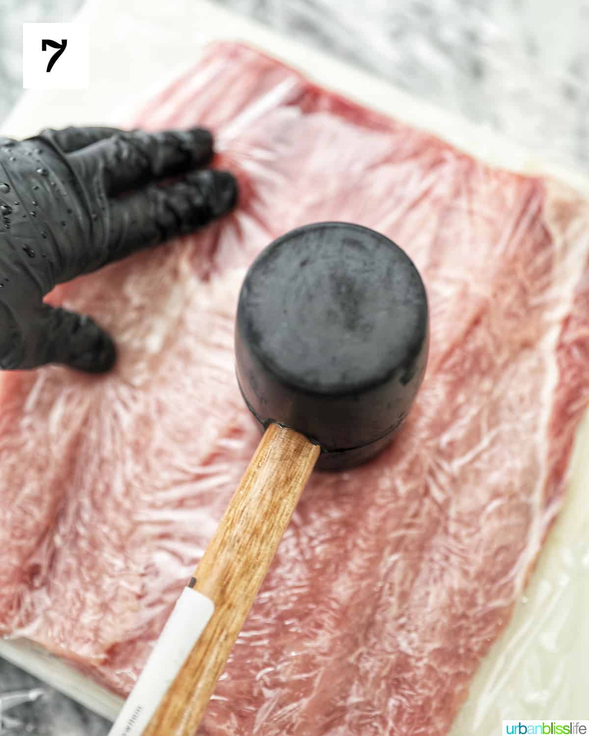 gloved hand using a meat mallet to tenderize and flatten the pork loin.