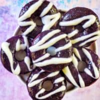 several ube donuts with ube glaze and white glaze drizzle on a purple and blue table.
