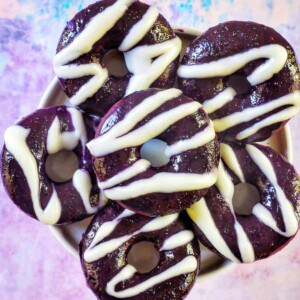 several ube donuts with ube glaze and white glaze drizzle on a purple and blue table.