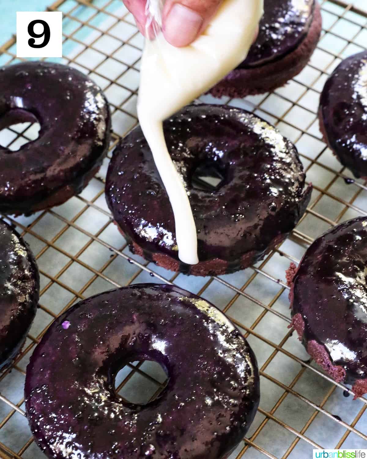 several ube donuts on a baking rack with a hand drizzling white glaze over donuts.