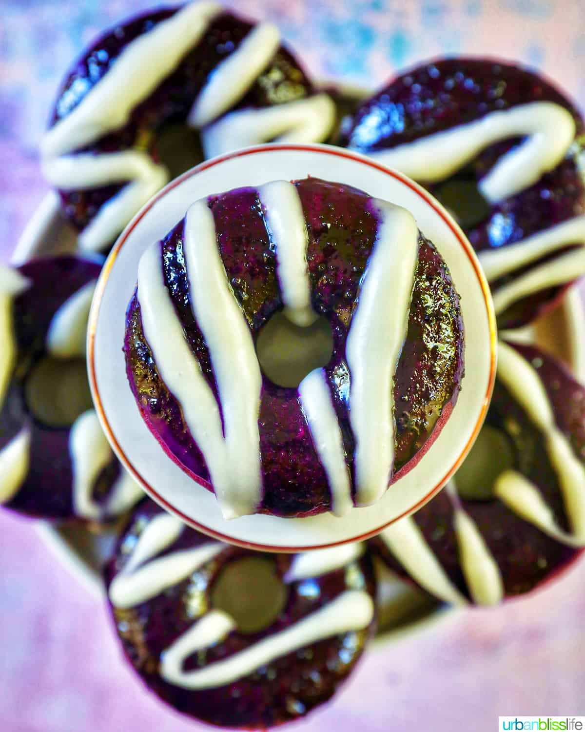 several ube donuts with ube glaze and white glaze drizzle, one on a pedestal in the middle.