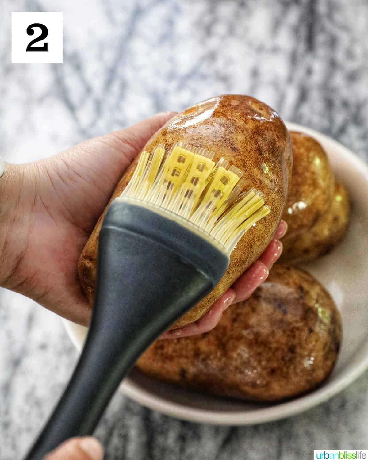 a hand holding a potato while the other hand brushes olive oil over the potato skin.