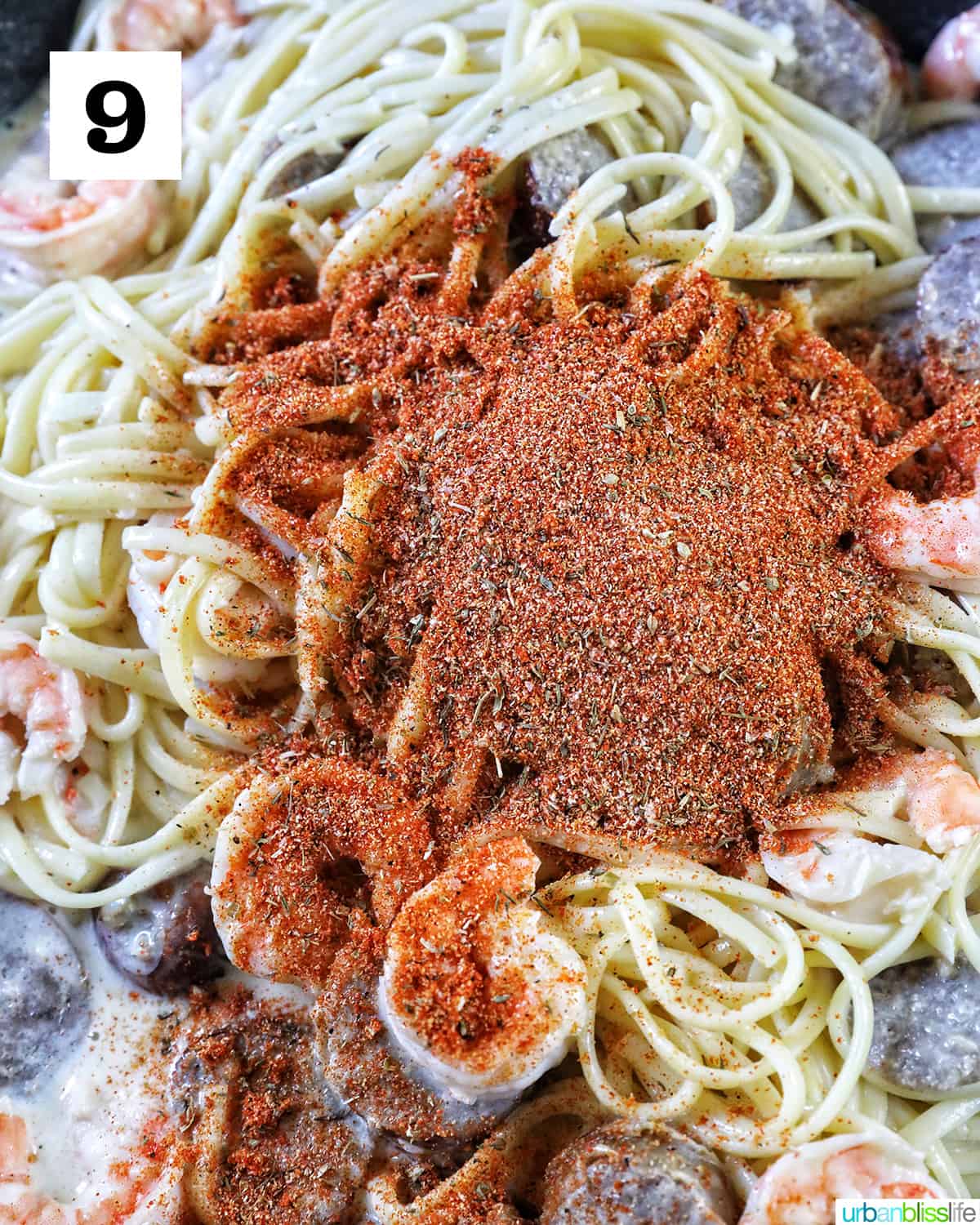 seasoning added to pasta with shrimp and sausage cooking in a skillet.