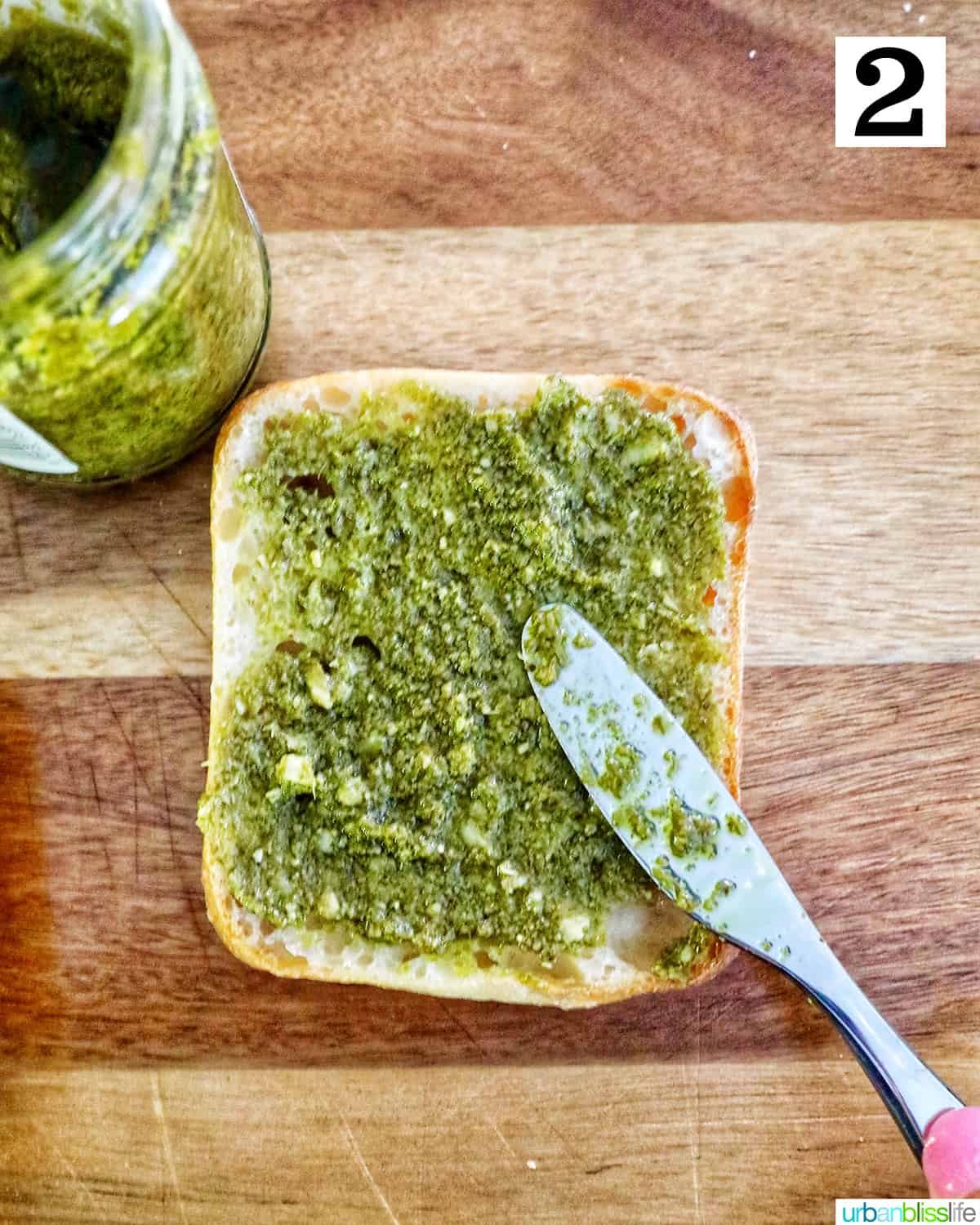 knife spreading pesto on top of a slice of ciabatta bread on a cutting board.