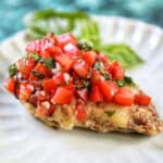 single bruschetta chicken on a white plate with basil leaves in the background.