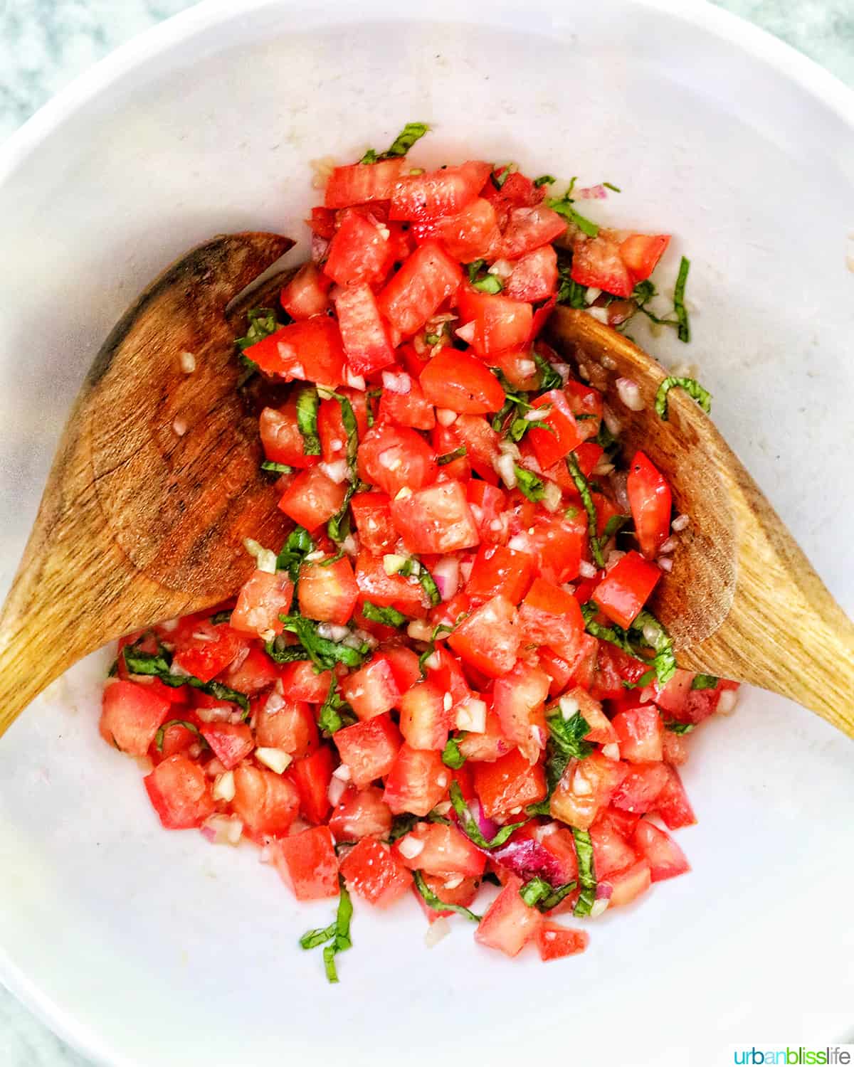 two wooden spoons tossing together bruschetta tomatoes, basil, and garlic.