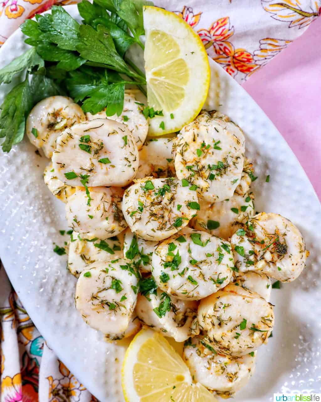 several cooked scallops topped with herbs and garlic, with lemon slices and parsley, on a white plate.