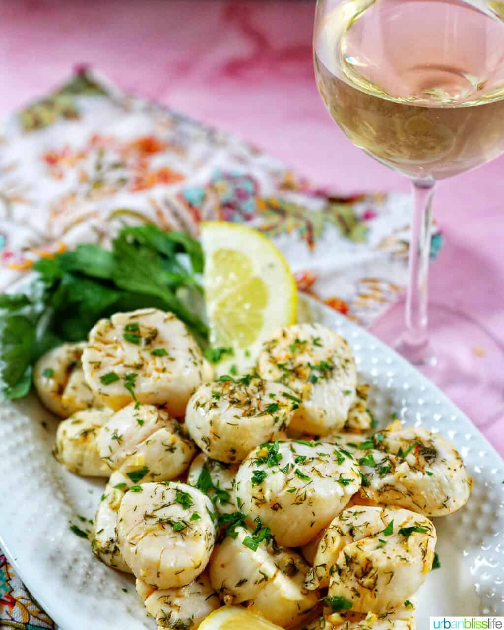 several cooked scallops topped with herbs and garlic, with lemon slices and parsley, on a white plate with a glass of white wine next to it.
