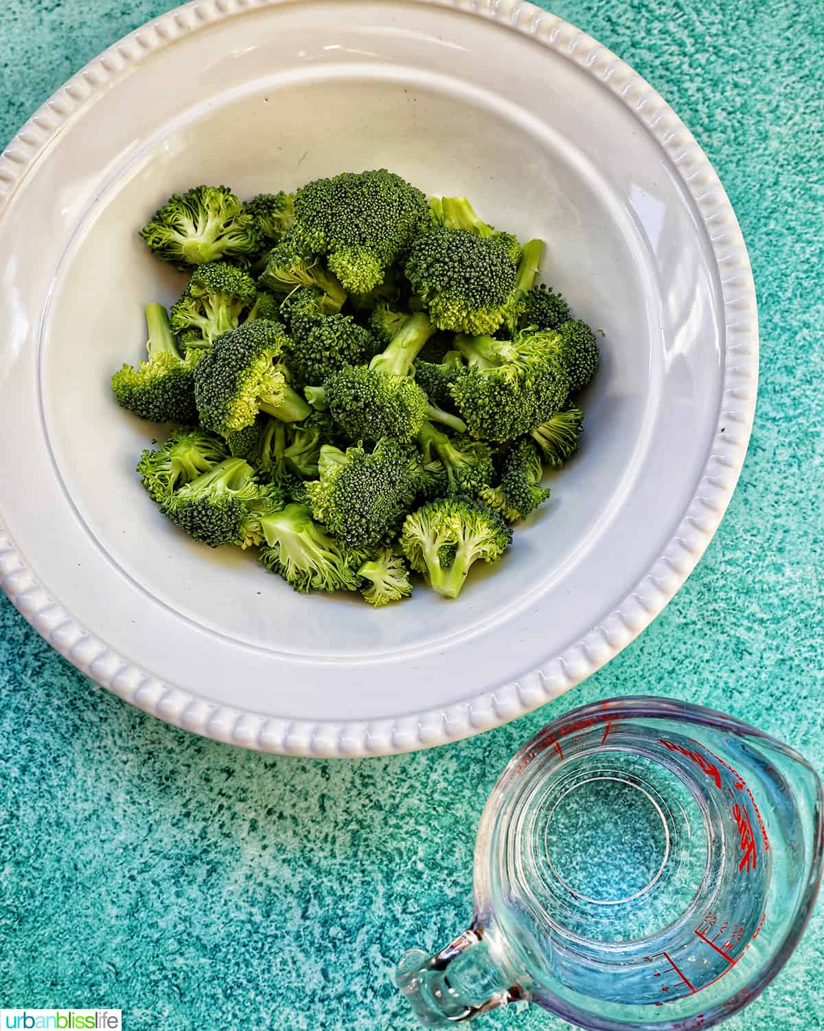 bowl of broccoli florets next to a cup of water on a blue table.
