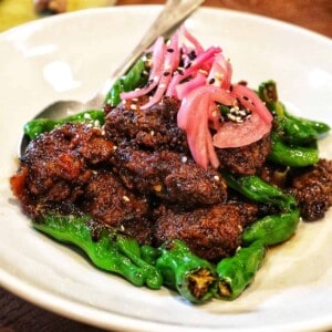 ginger beef with pickled onions and shishito peppers on a white plate.