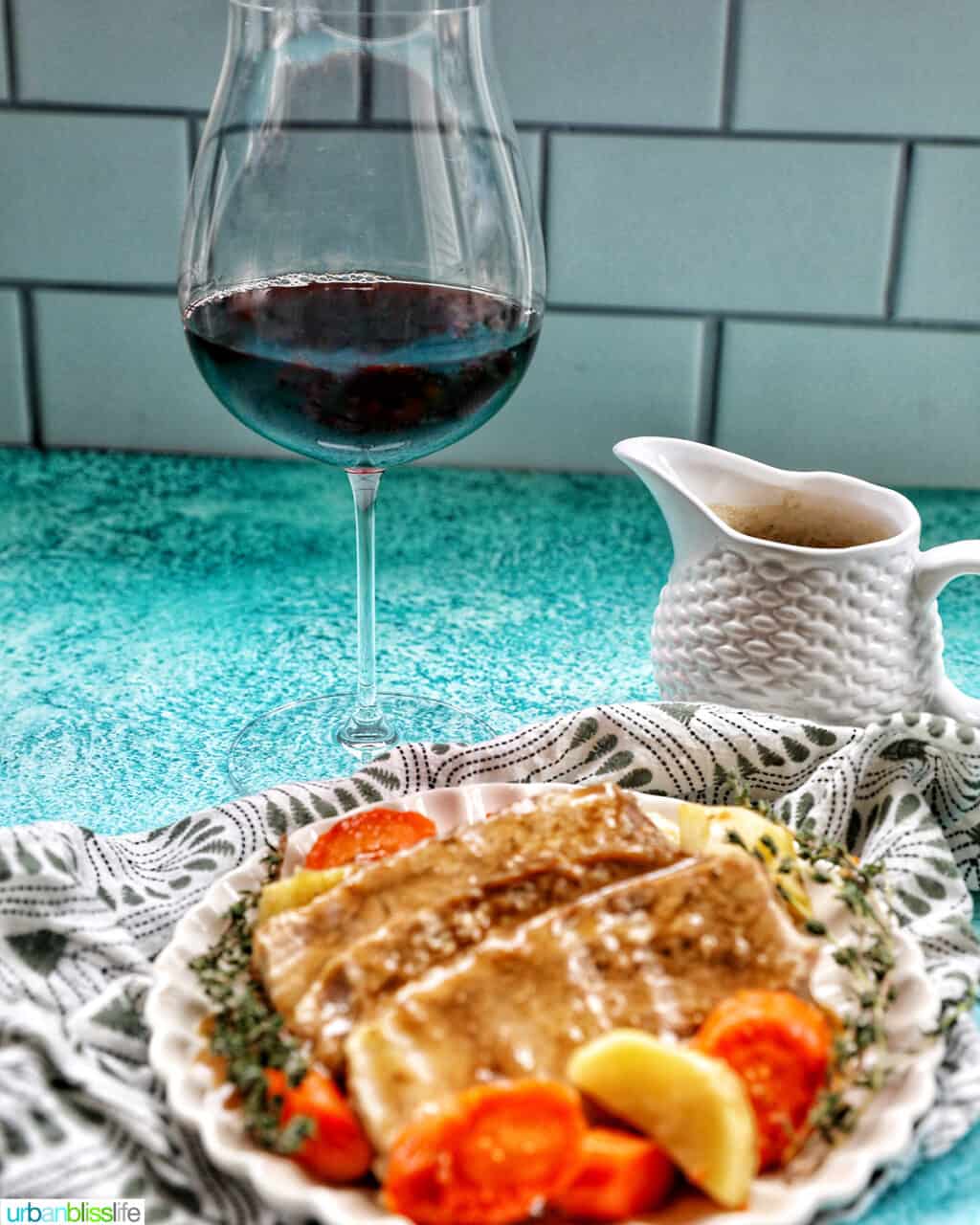 Glass of Pinot Noir red wine behind a plate with sliced pork roast, carrots, potatoes, and a gravy boat with gravy on the side.