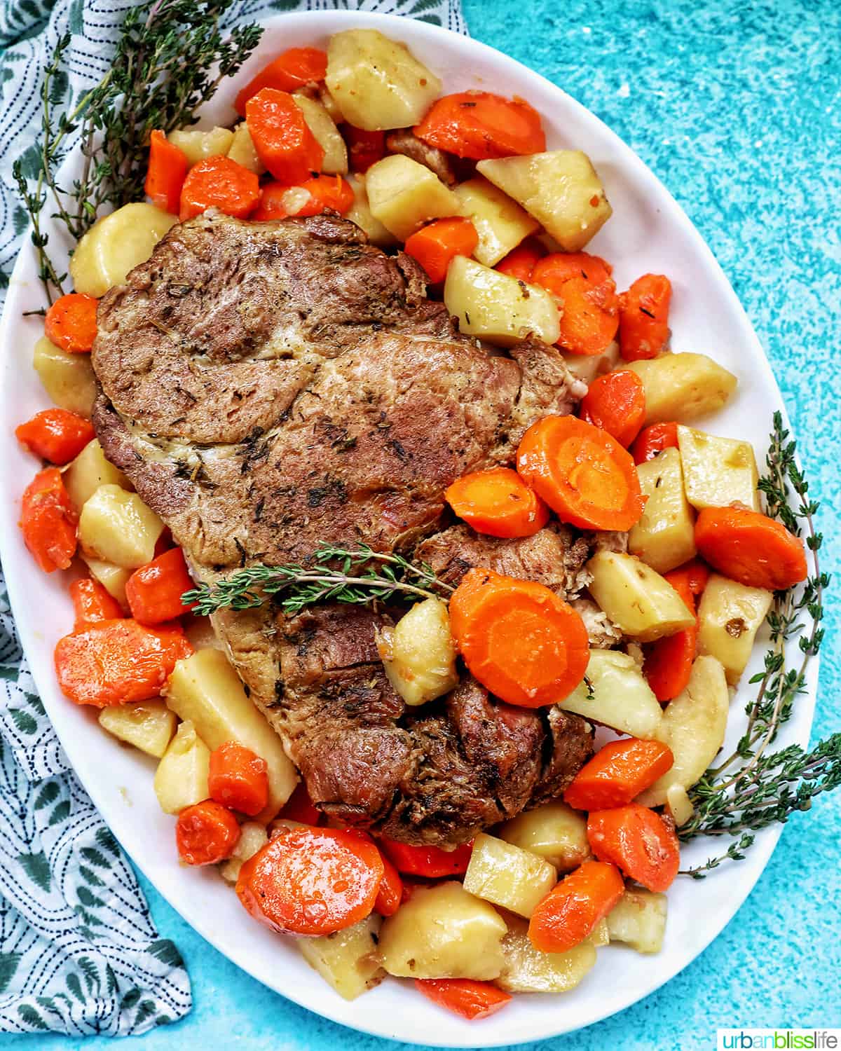 Instant Pot pork roast surrounded by chopped carrots, potatoes, and herbs on a white serving platter on a blue table.