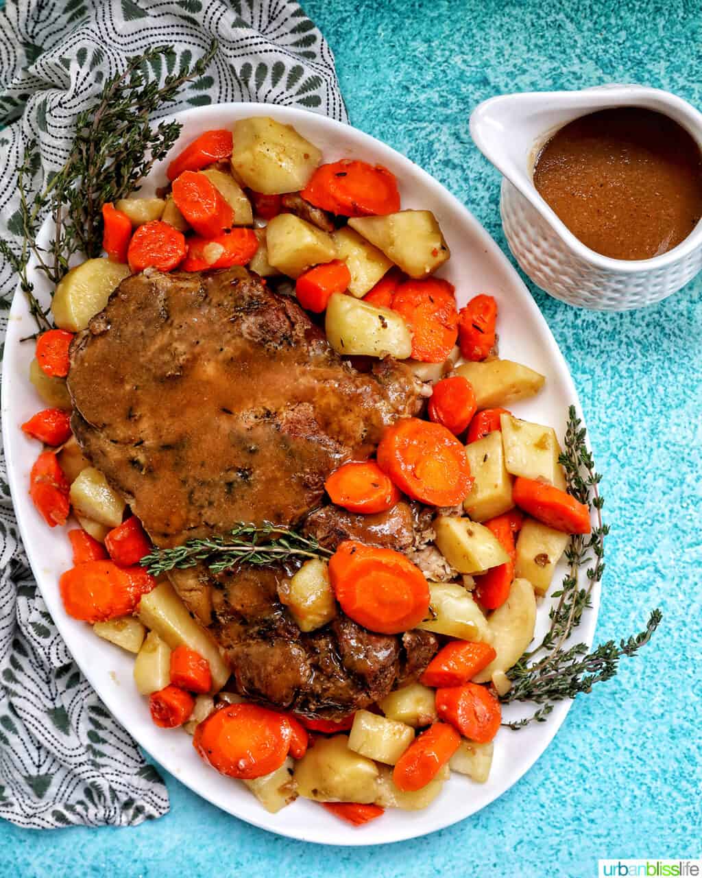 Instant Pot pork roast surrounded by chopped carrots, potatoes, and herbs on a white serving platter with a side of gravy in a gravy boat on a blue table.