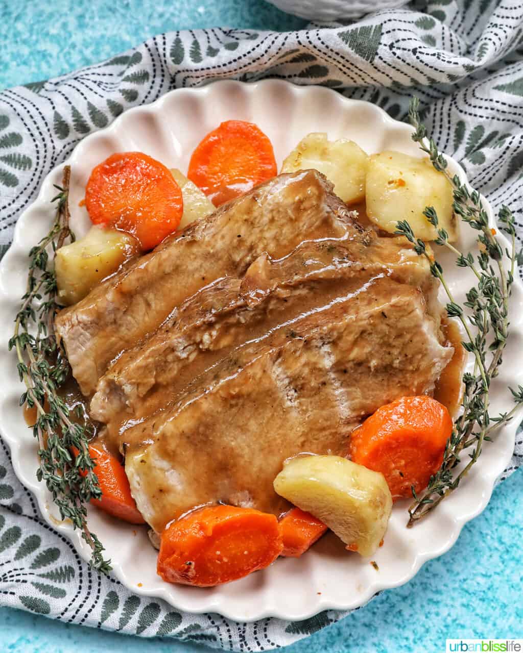 sliced pork roast with sliced carrots and potatoes, a garnish of herbs, on a white plate on a blue table.