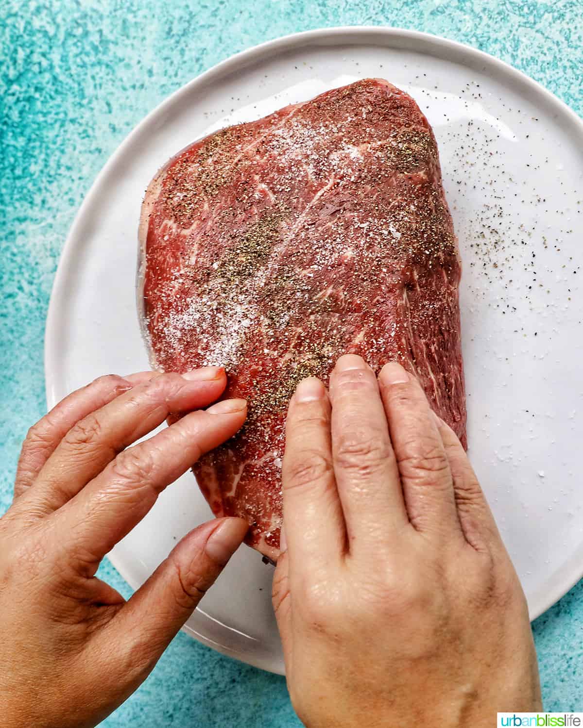two hands seasoning sirloin tip roast with salt and pepper on a white plate.