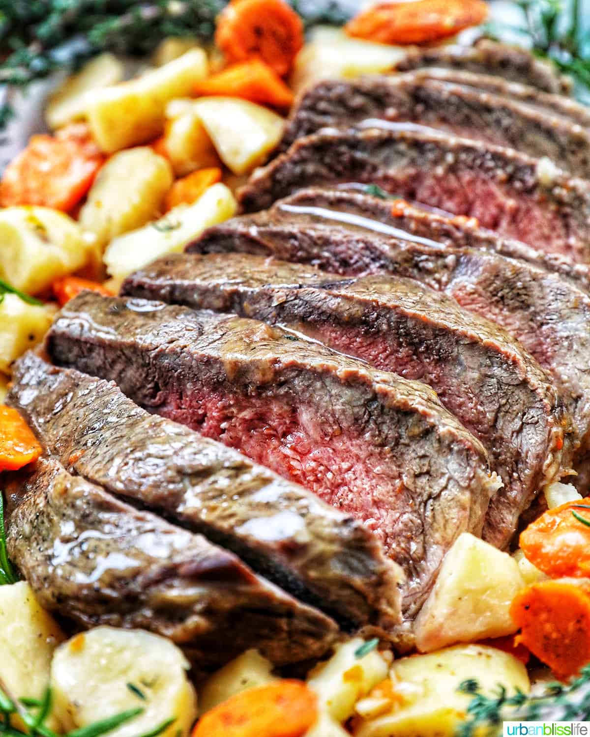 Sliced sirloin tip roast surrounded by carrots, potatoes, and herbs.