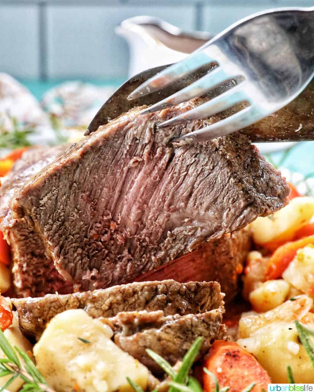 Large fork and knife holding up a slice of sirloin tip roast with potatoes and carrots in front.