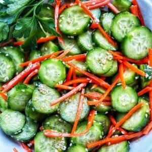 Bowl of sliced cucumbers, julienned carrots, sesame seeds, and herbs on the side.
