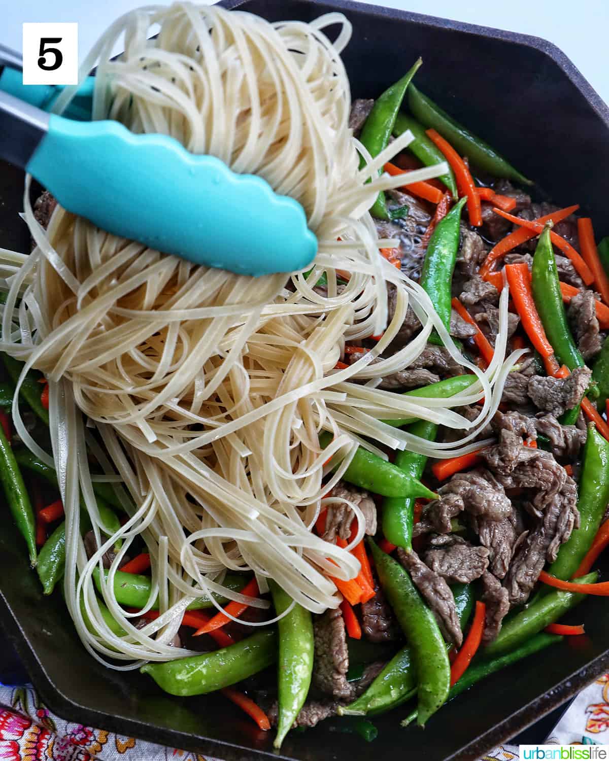 blue tongs holding up beef pad thai noodles with vegetables in a cast iron skillet.