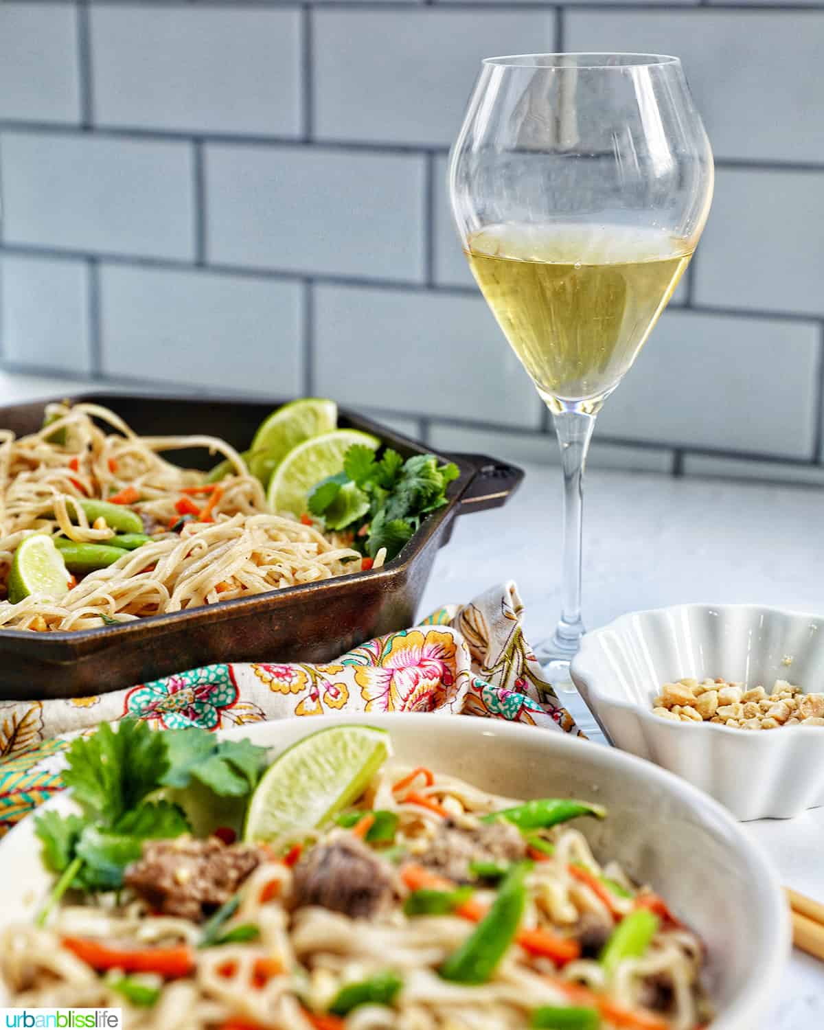 bowl of beef pad thai noodles with vegetables with glass of white wine and skillet of more pad thai.
