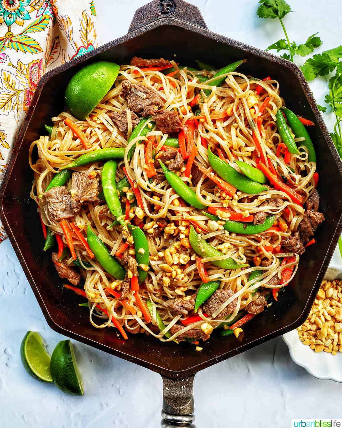 beef pad thai noodles with vegetables in a Finex cast iron skillet with side of lime wedges and chopped peanuts.