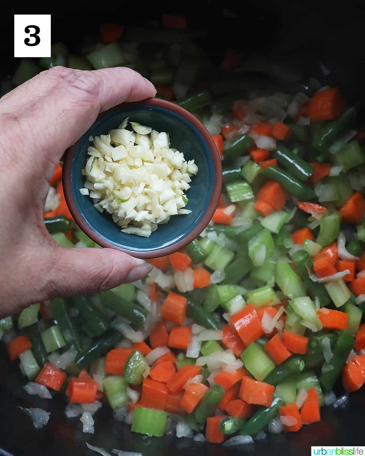 hand holding a bowl of minced garlic over vegetables.
