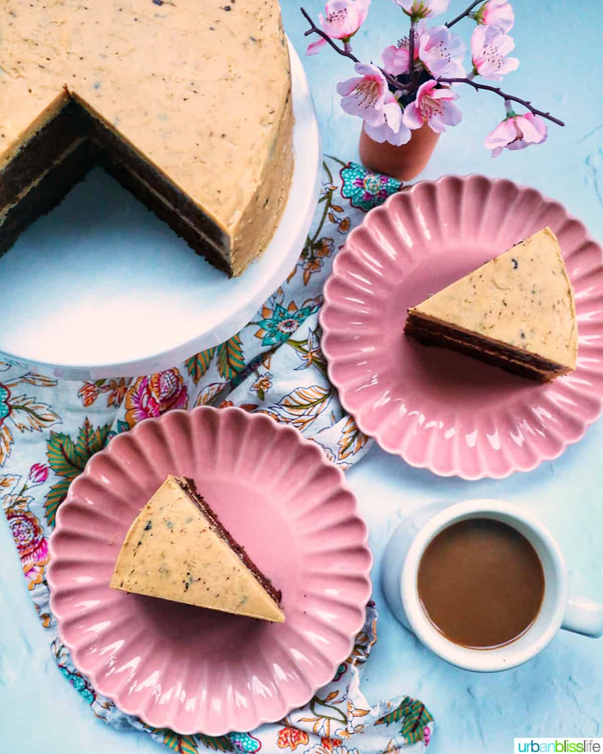 chocolate coffee cake on a pedestal with plates of cake slices, cup of coffee, and pink flowers.