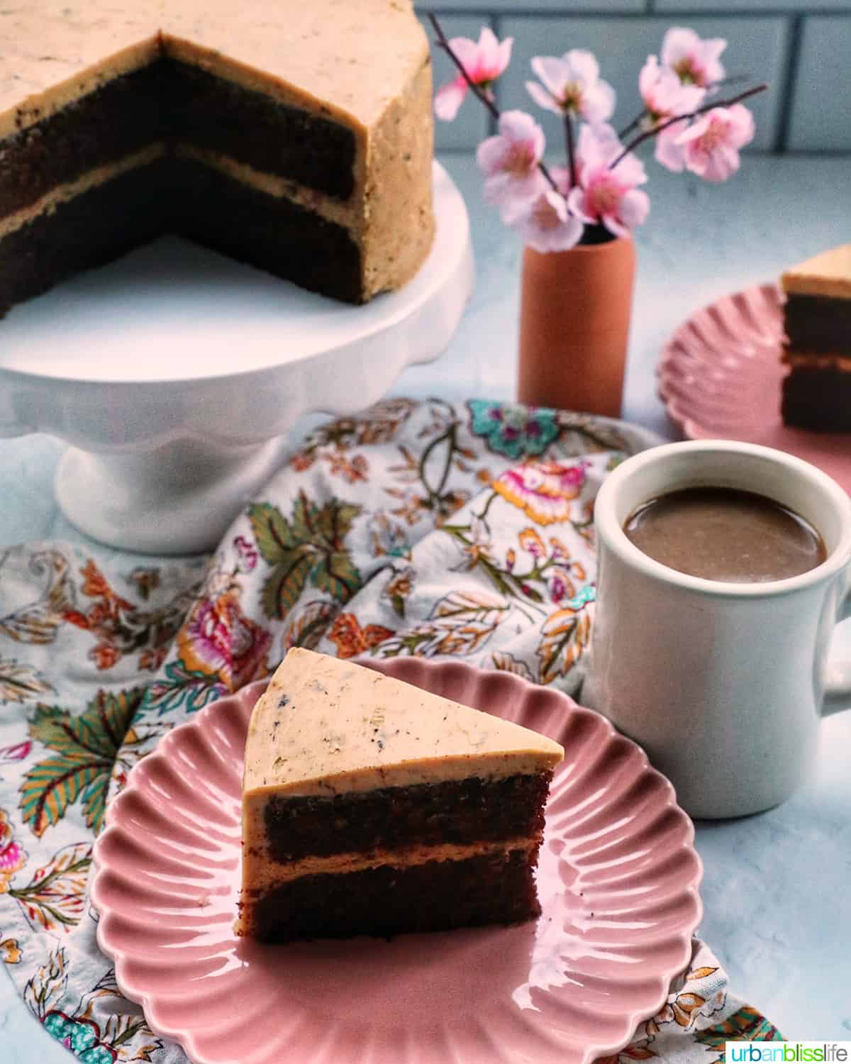 chocolate coffee cake on a pedestal with plates of cake slices, cup of coffee, and pink flowers.