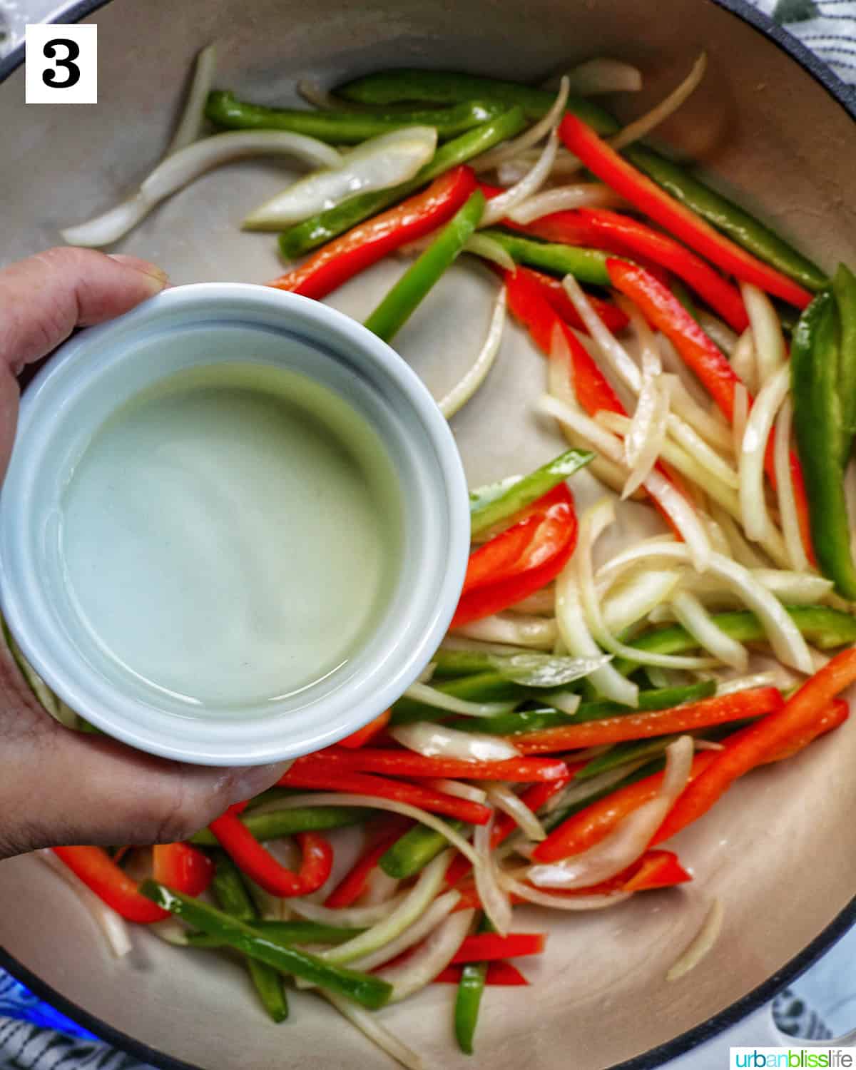 Hand holding bowl of white wine over sliced onions, red peppers, and green peppers in a saucepan.