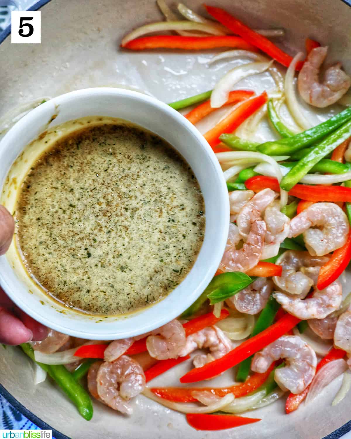 Hand holding bowl of sauce over sliced onions, red peppers, and green peppers in a saucepan.