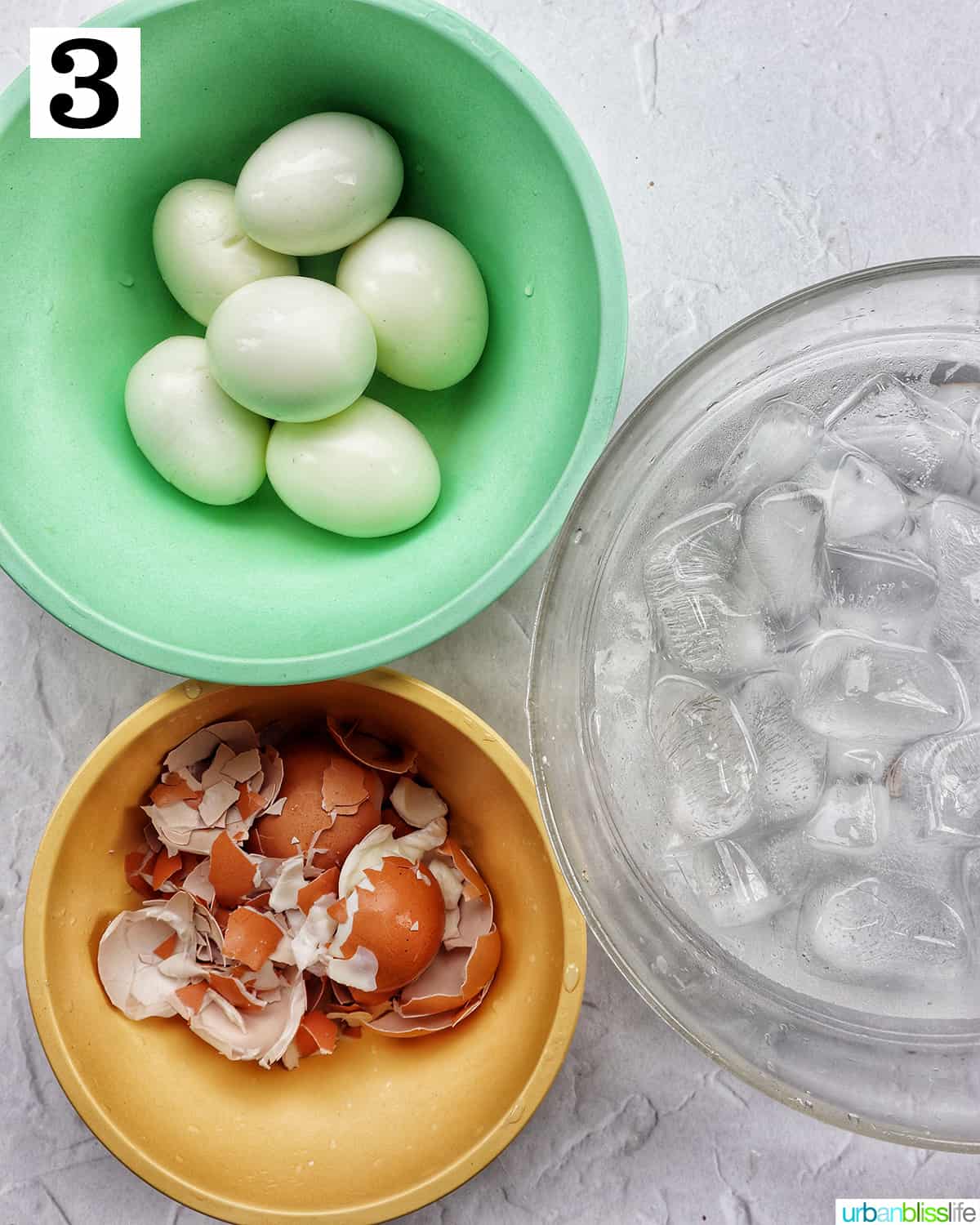green bowl with peeled hardboiled eggs, yellow bowl with peeled eggshells, and clear bowl with ice water bath.