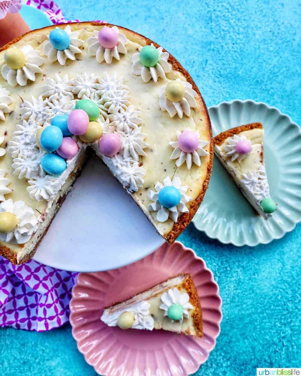 easter egg cheesecake with mini chocolate eggs and whipped cream, with two slices on plates next to the main cake.