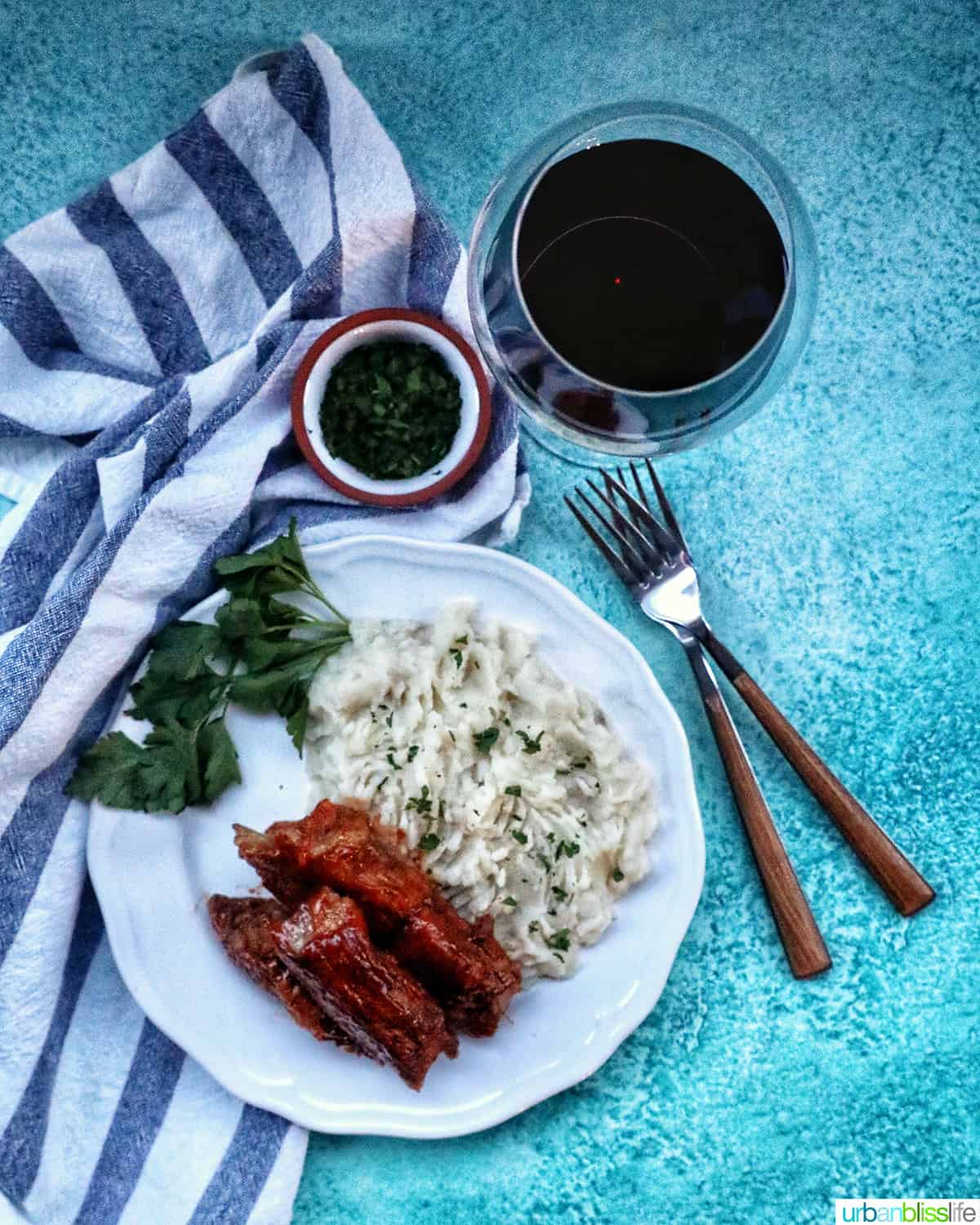 country style ribs and mashed potatoes on a white plate with two forks and glass of red wine.