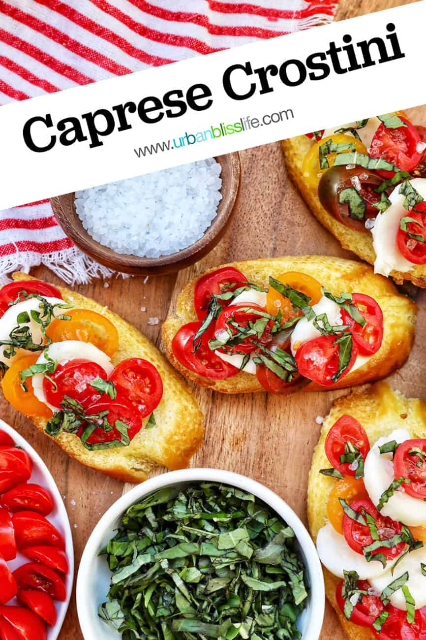 crostini with tomatoes, mozzarella, basil surrounded by plates of the same toppings and a bowl of sea salt with title text that reads Caprese Crostini.