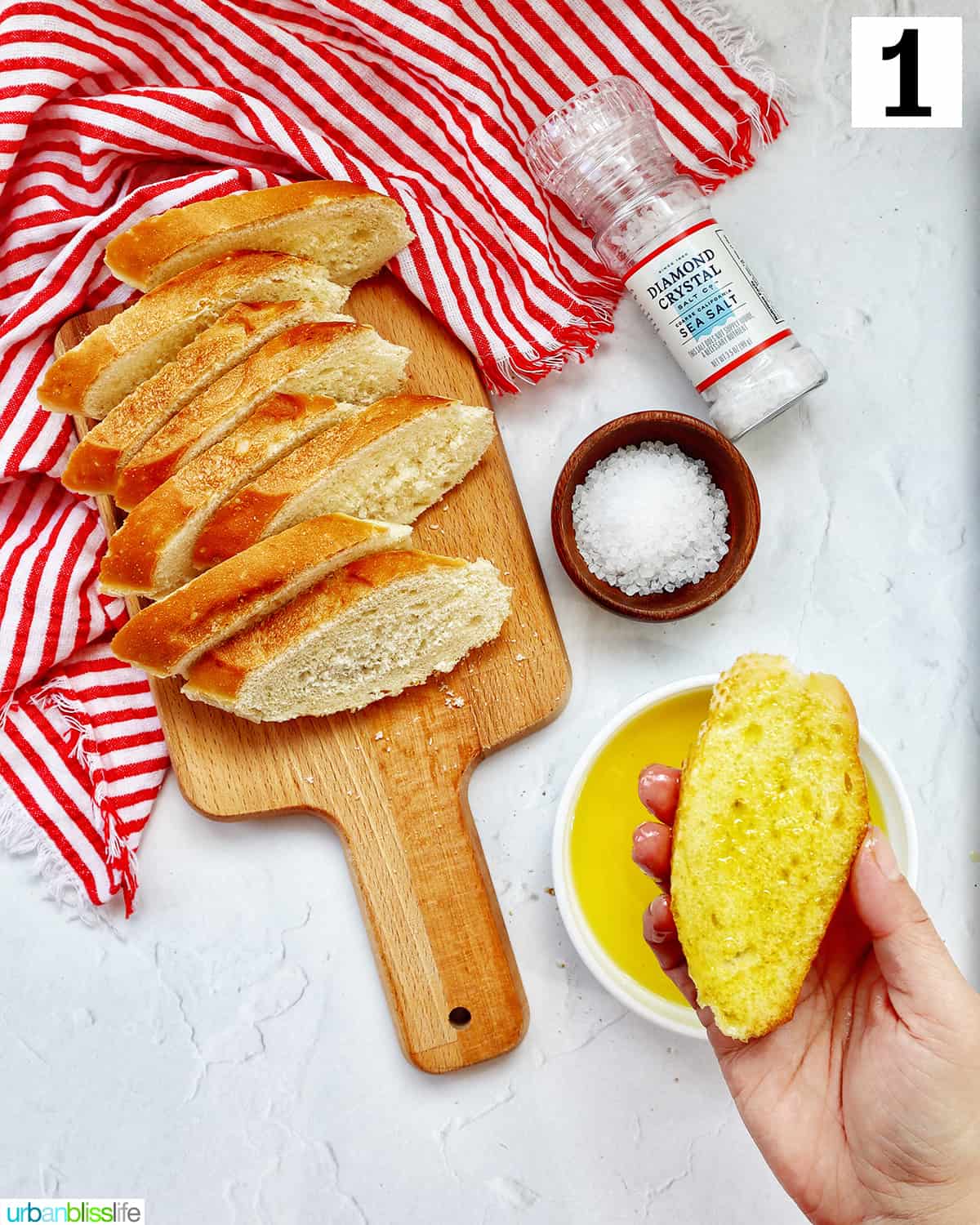 hand dipping a crostini into a bowl of olive oil with crostini slices on a wooden cutting board on red and white towell with bowl and shaker of sea salt.