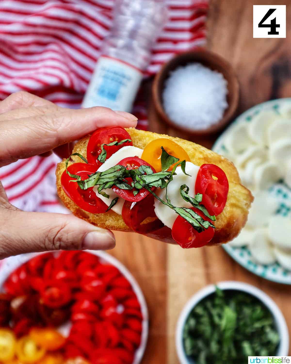hand holding a crostini with tomatoes, mozzarella, basil above a table with plates of the same toppings and a bowl of sea salt.