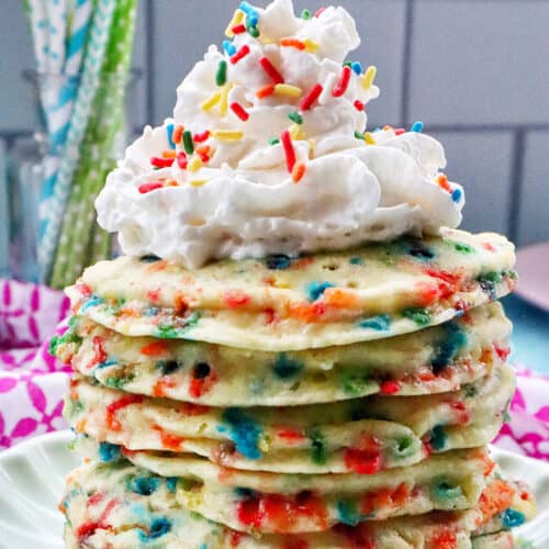 huge stack of funfetti pancakes with whipped cream and sprinkles on a scalloped green plate.