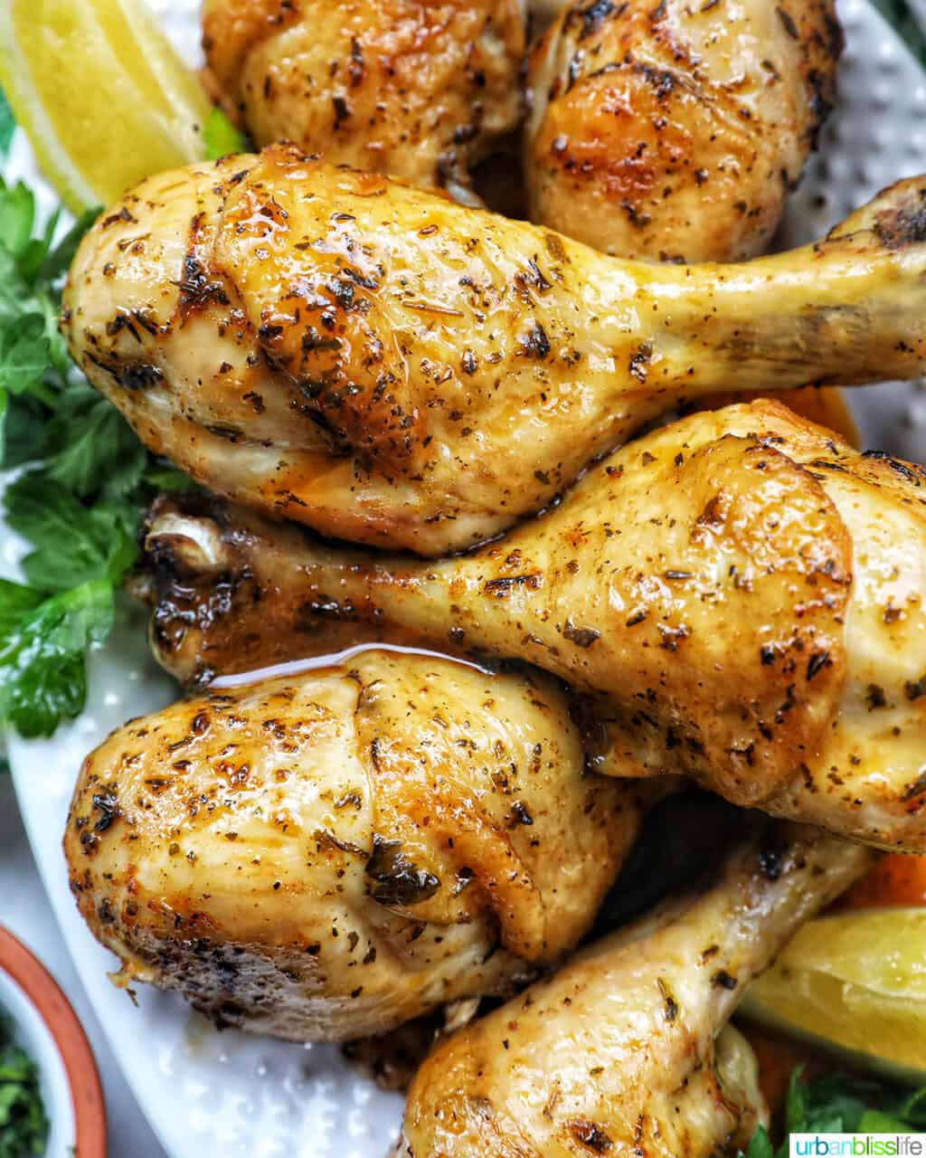 Stack of chicken drumsticks with seasoning and garnished with parsley and side of lemon wedges on bed of herbs.