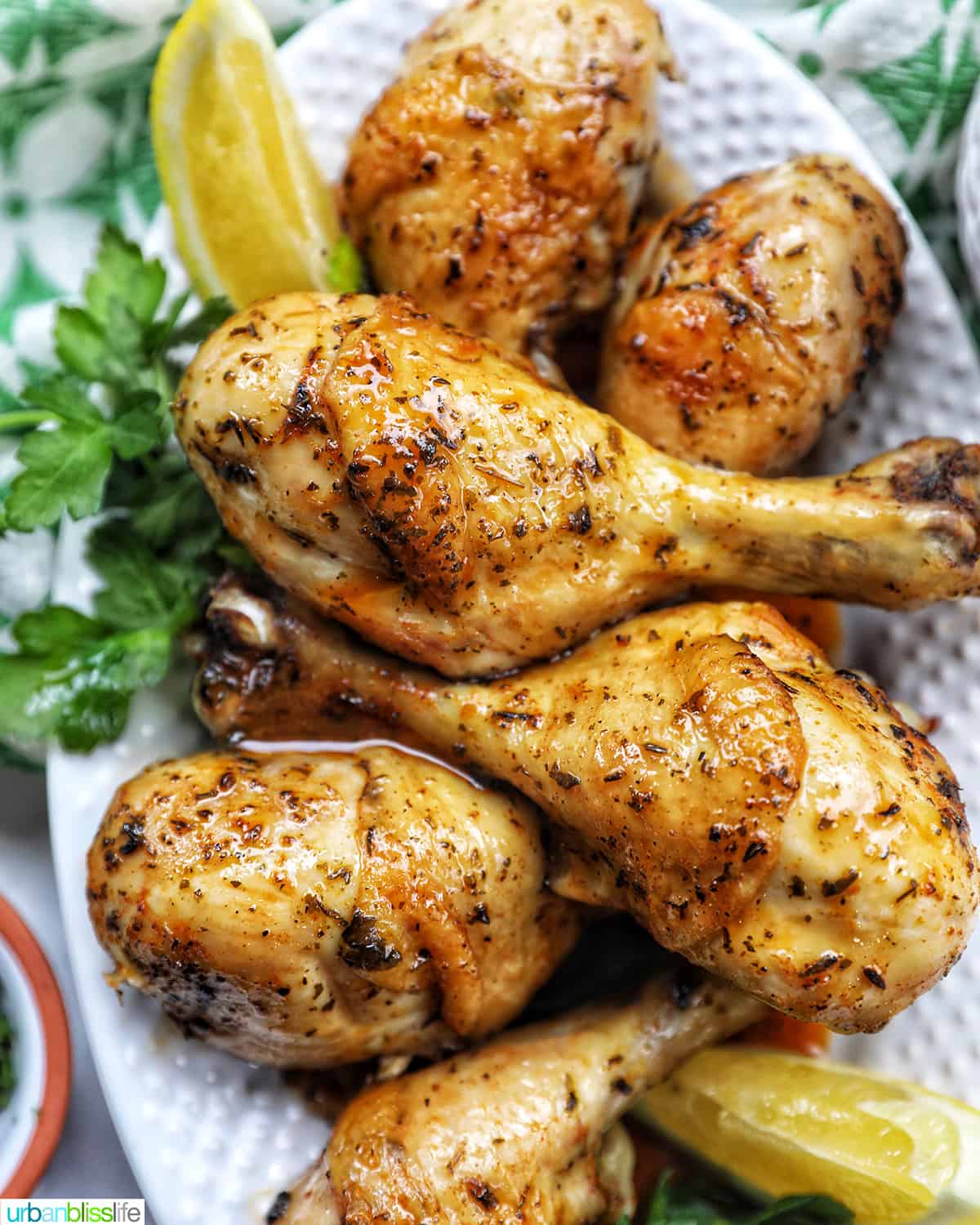 Stack of chicken drumsticks with seasoning and garnished with parsley and side of lemon wedges on bed of herbs.