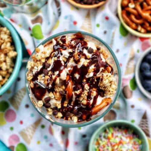ice cream parfait topped with granola, chocolate syrup, mini chocolate chips on a colorful kitchen towel surrounded by sides of more toppings.