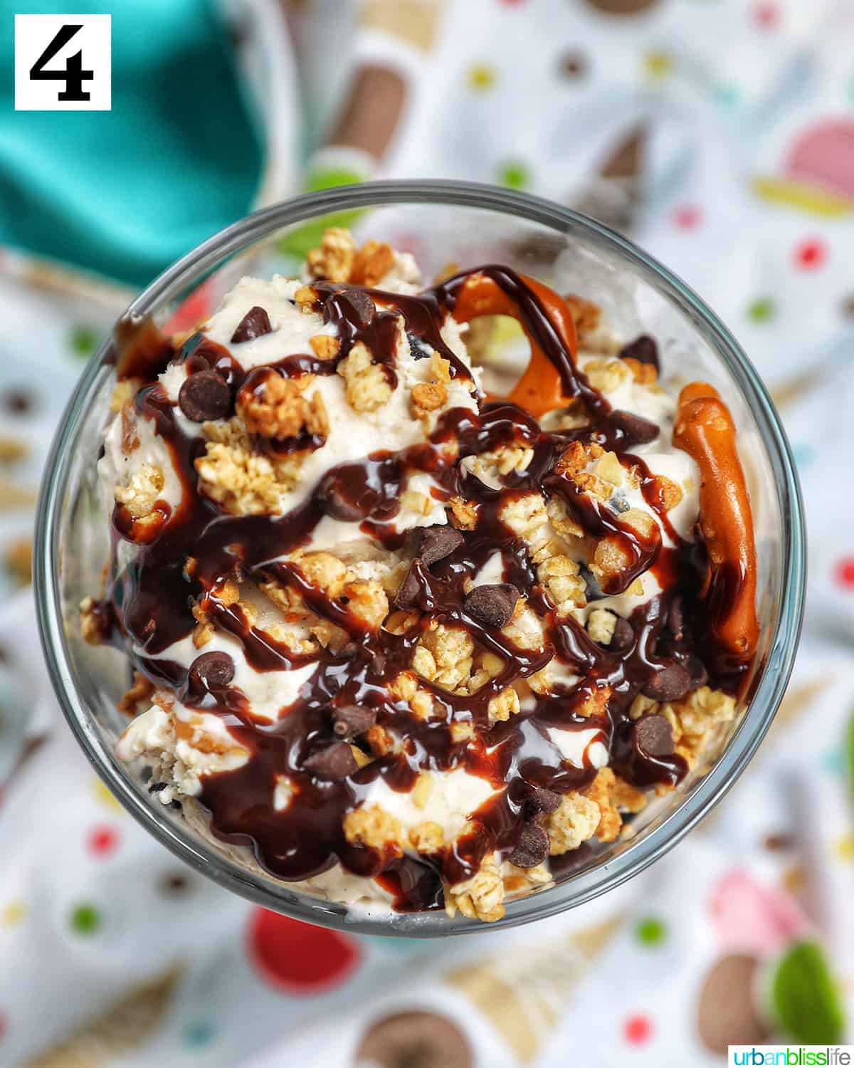 ice cream parfait topped with granola, chocolate syrup, mini chocolate chips on a colorful kitchen towel.