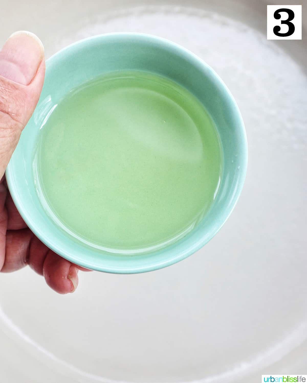 hand holding a small bowl of olive oil over a pan.