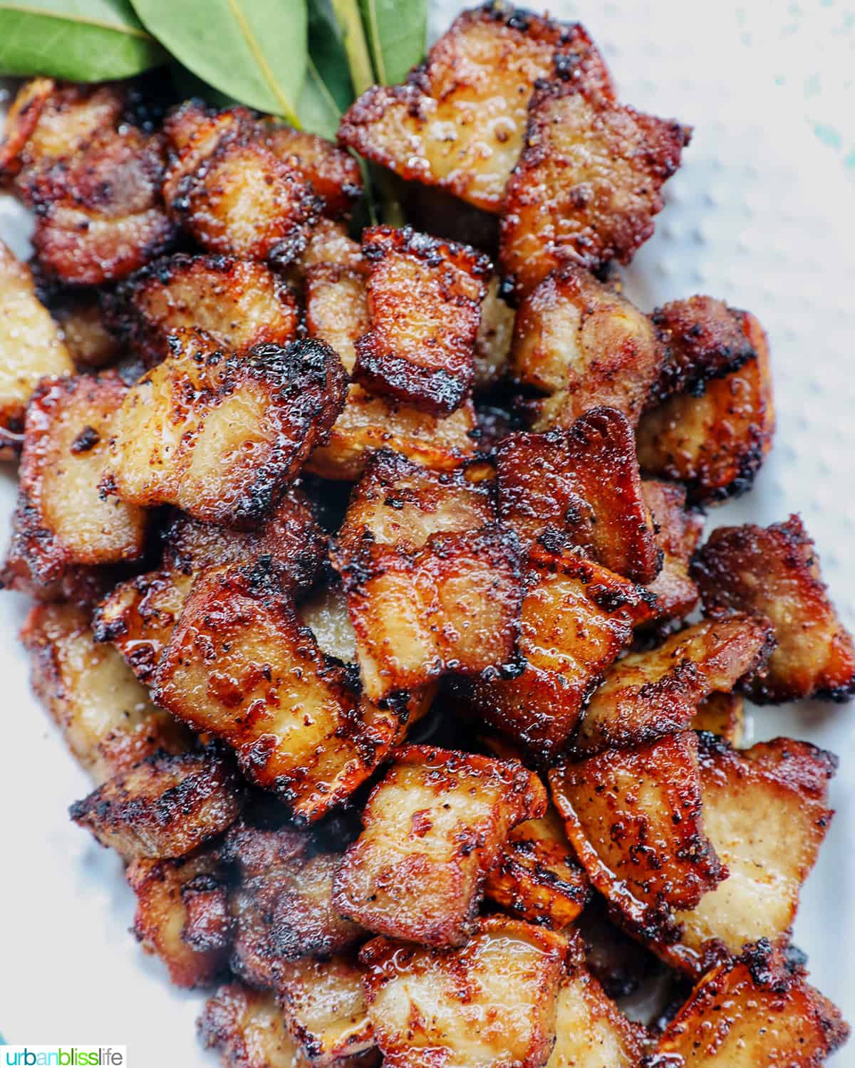 pile of air fryer pork belly bites on a white serving platter with bay leaves on a blue background.