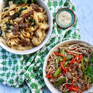 pad see ew in a white bowl above pad thai in a white bowl, both on a green patterned napkin with side of sesame seeds.
