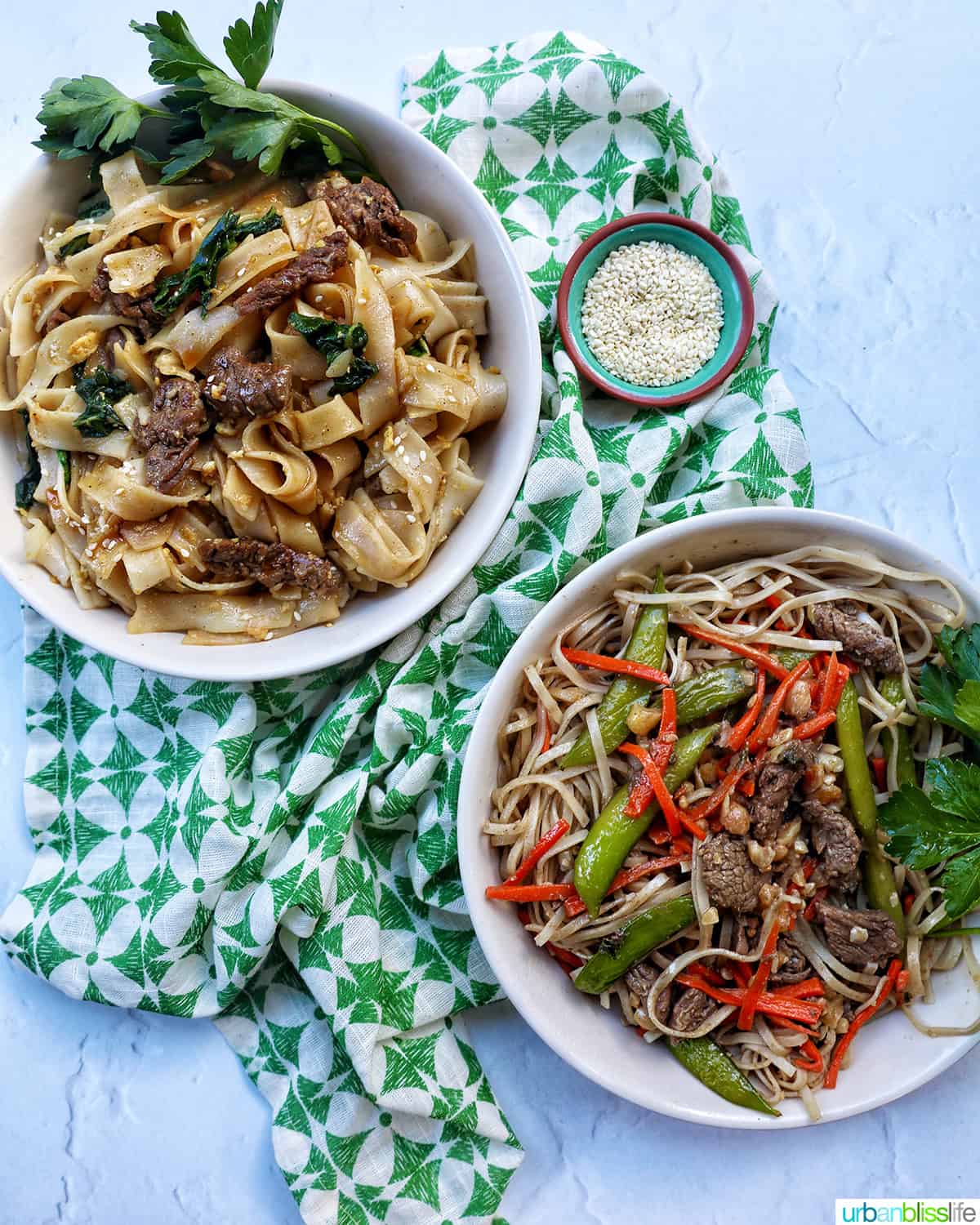 pad see ew in a white bowl above pad thai in a white bowl, both on a green patterned napkin with side of sesame seeds.