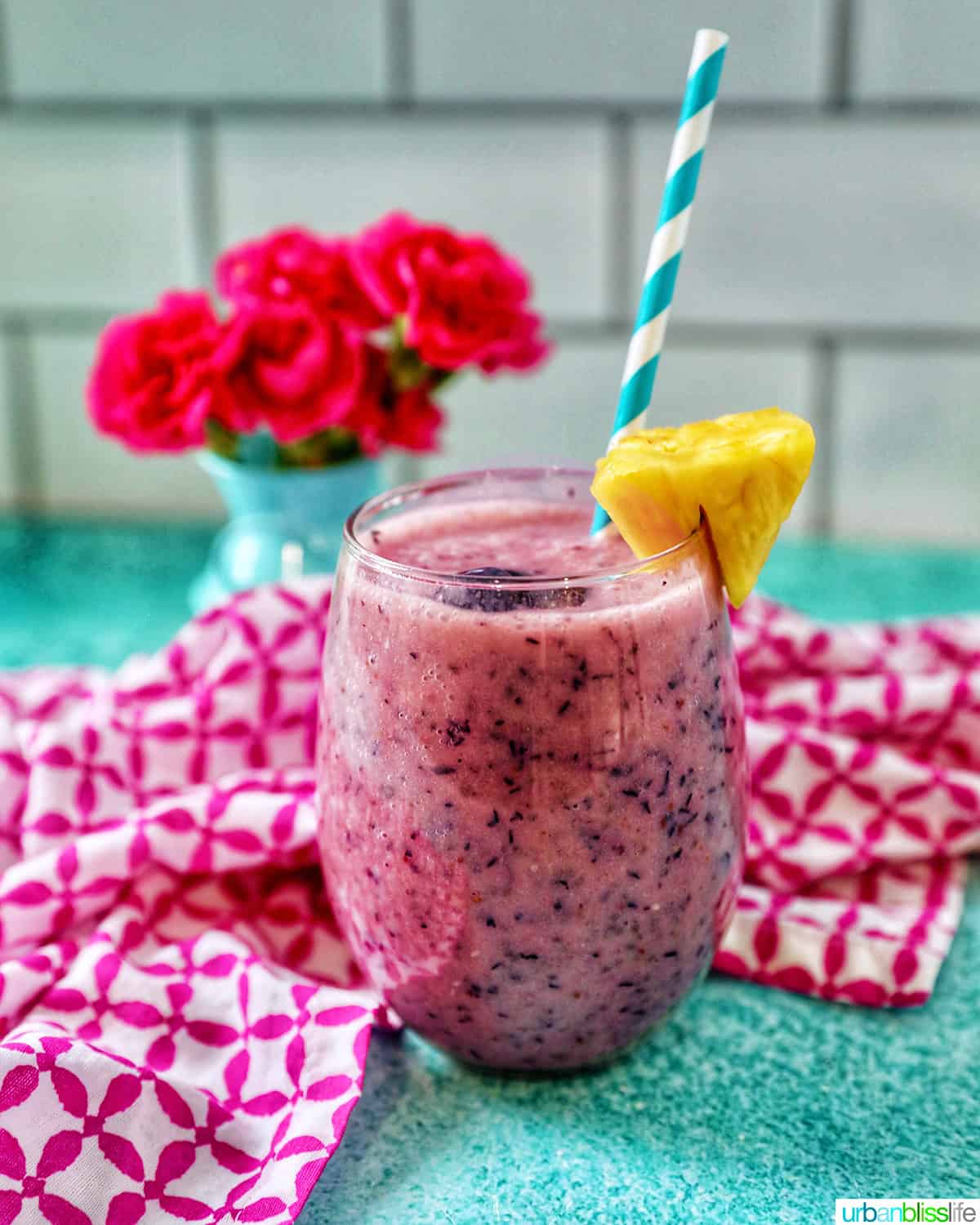 glass of Blueberry Pineapple Smoothie with pineapple wedge garnish, blue and white stripe straw, and pink and white napkin.