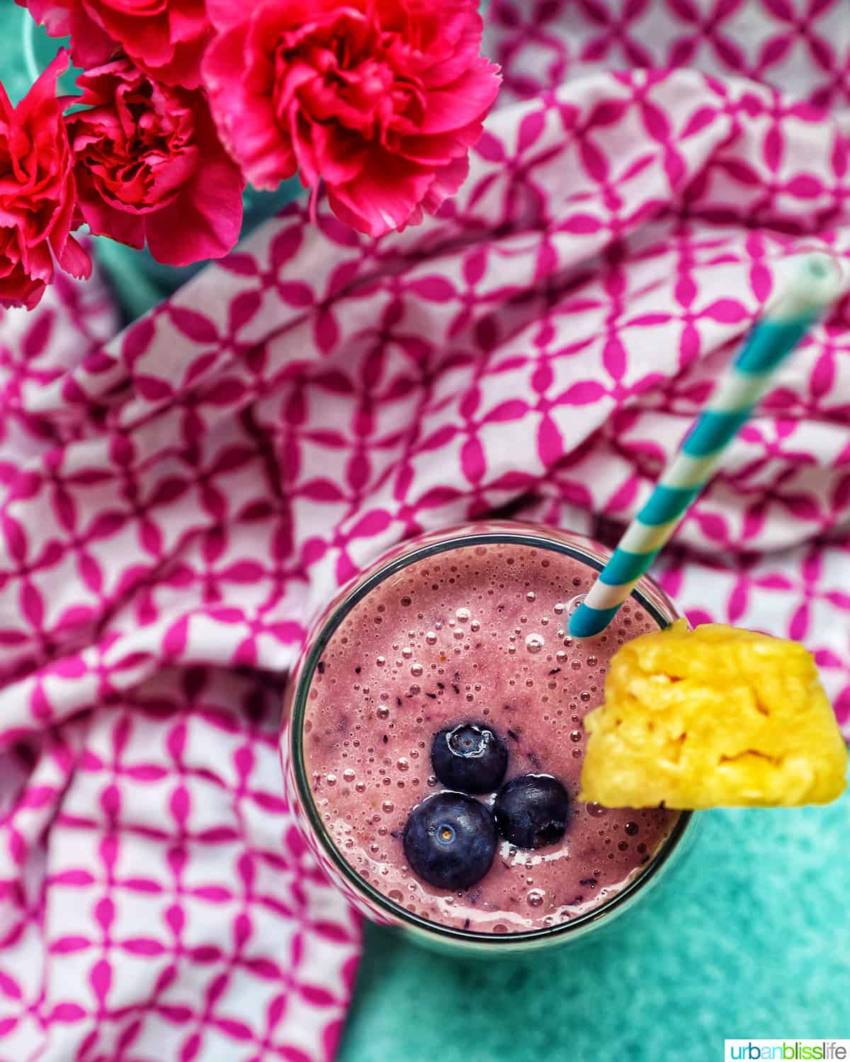 glass of Blueberry Pineapple Smoothie with pineapple wedge garnish, blue and white stripe straw, and pink and white napkin.