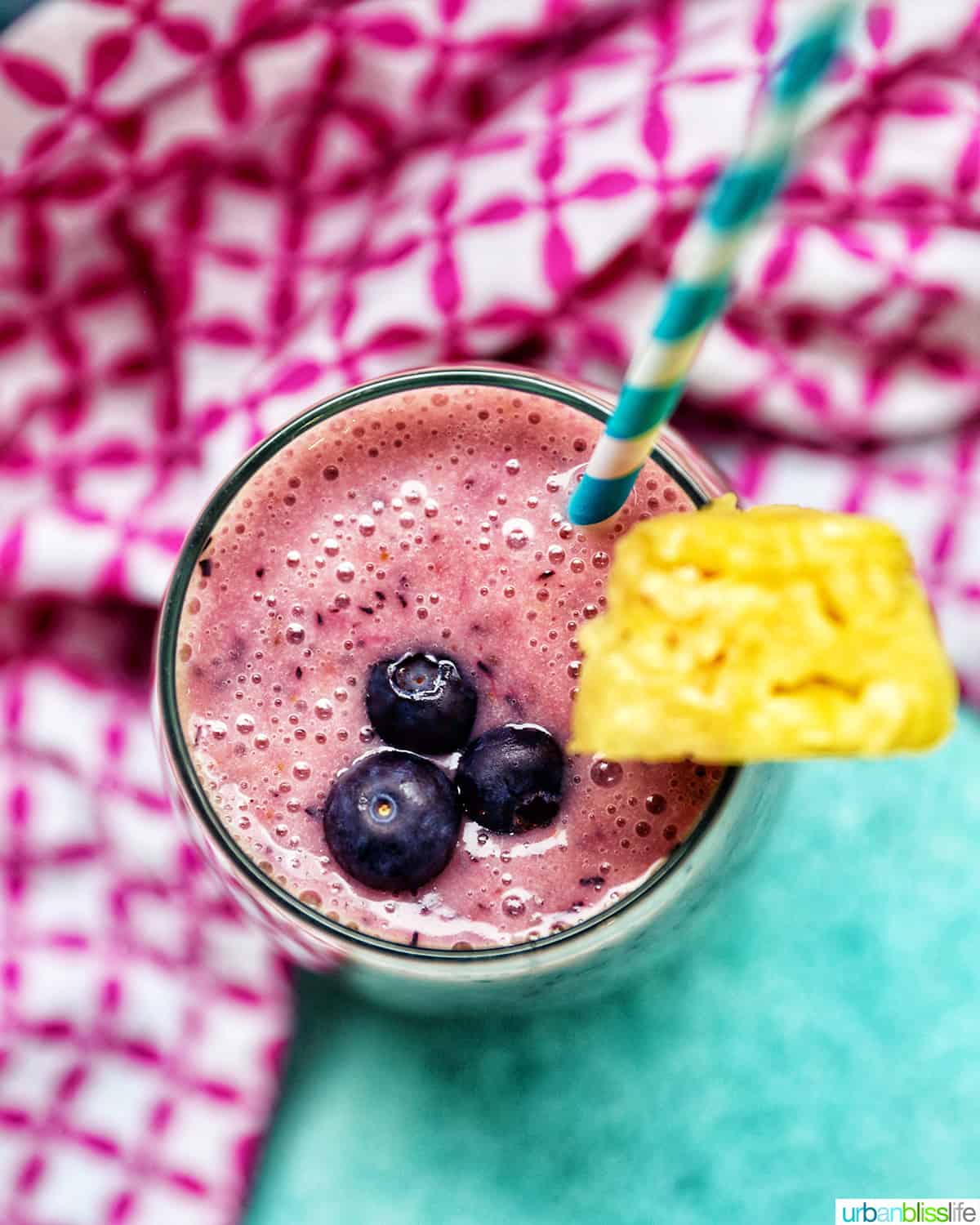 glass of Blueberry Pineapple Smoothie with pineapple wedge garnish, blue and white stripe straw, and pink and white napkin.