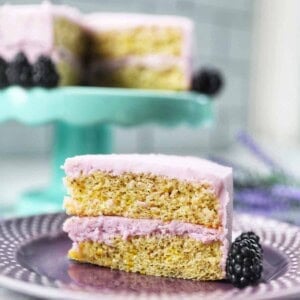 slice of earl grey lavender cake on a purple plate with blackberries with cake on an aqua cake pedestal in the background.