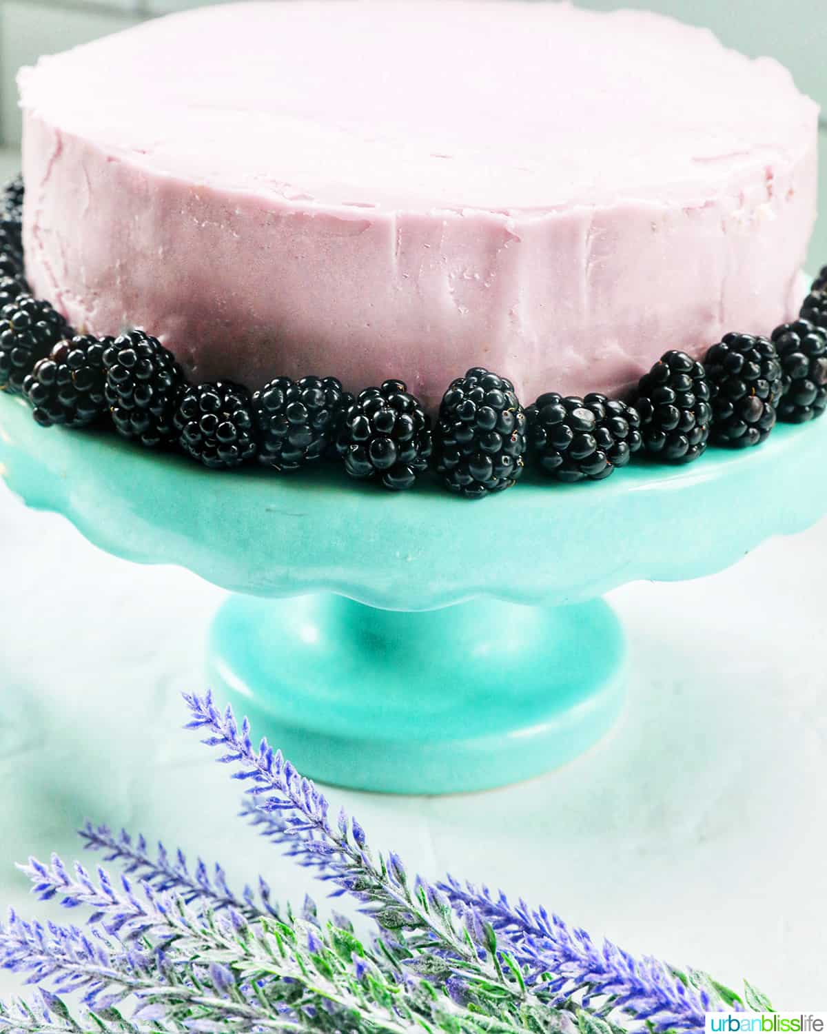 slice of earl grey lavender cake on a purple plate with blackberries with cake on an aqua cake pedestal in the background.